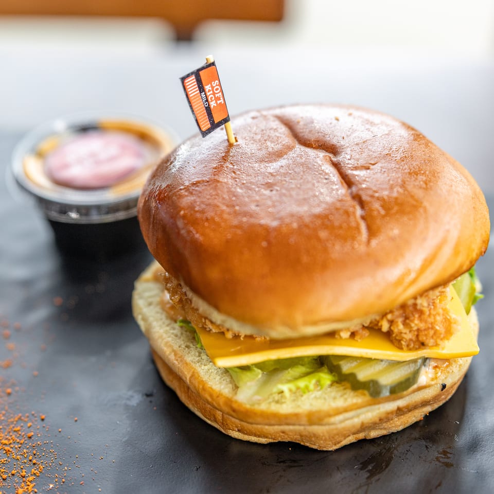 Close-up of a crispy chicken burger with cheddar, lettuce, pickles, and sauce, topped with a soft-pick flag.