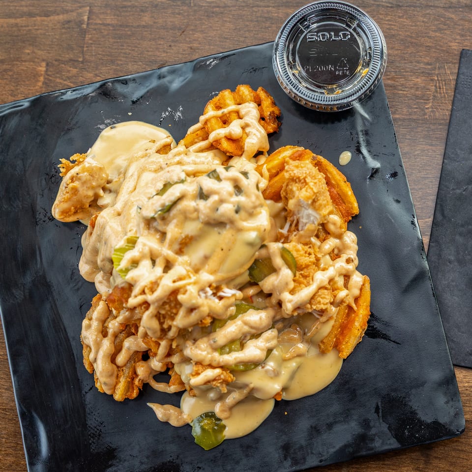 Crispy fried chicken and waffle fries topped with creamy sauces and pickles on a black plate.