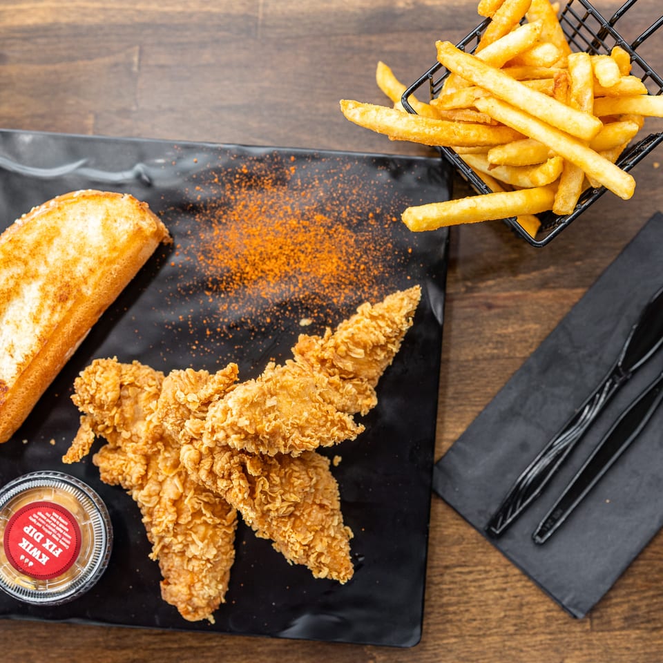 Crispy fried chicken tenders with toasted bread, French fries, dipping sauce, and black utensils arranged on a wooden table.