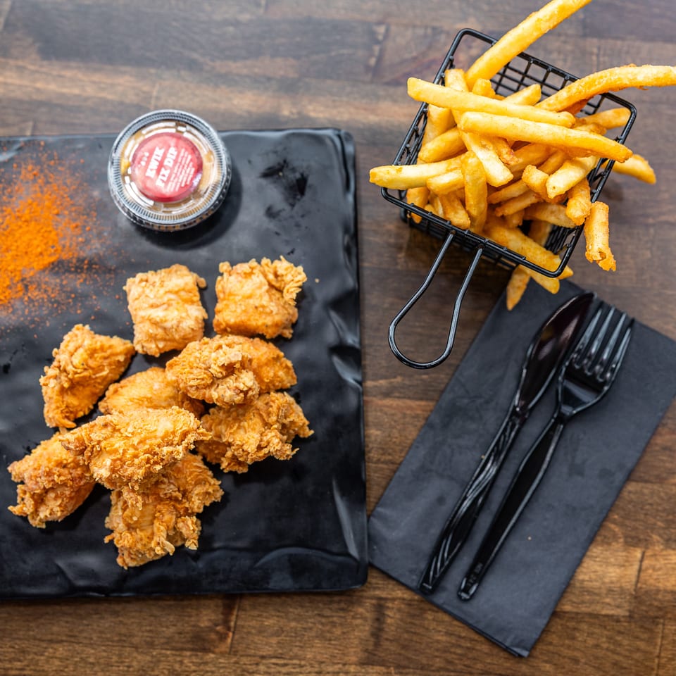 Overhead view of crispy fried chicken, golden fries in a wire basket, dipping sauce, and black utensils on a wooden table.