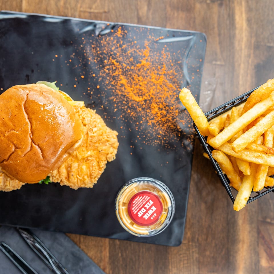 Crispy fried chicken sandwich with lettuce, French fries, dipping sauce, and orange seasoning on a black serving board.